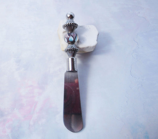 The Handcrafted Dip Spreader Knife – Glass Beads, Modern Entertaining features a silver-detailed glass bead handle and rests on white stone against a pastel background—an ideal accent for stylish gatherings.
