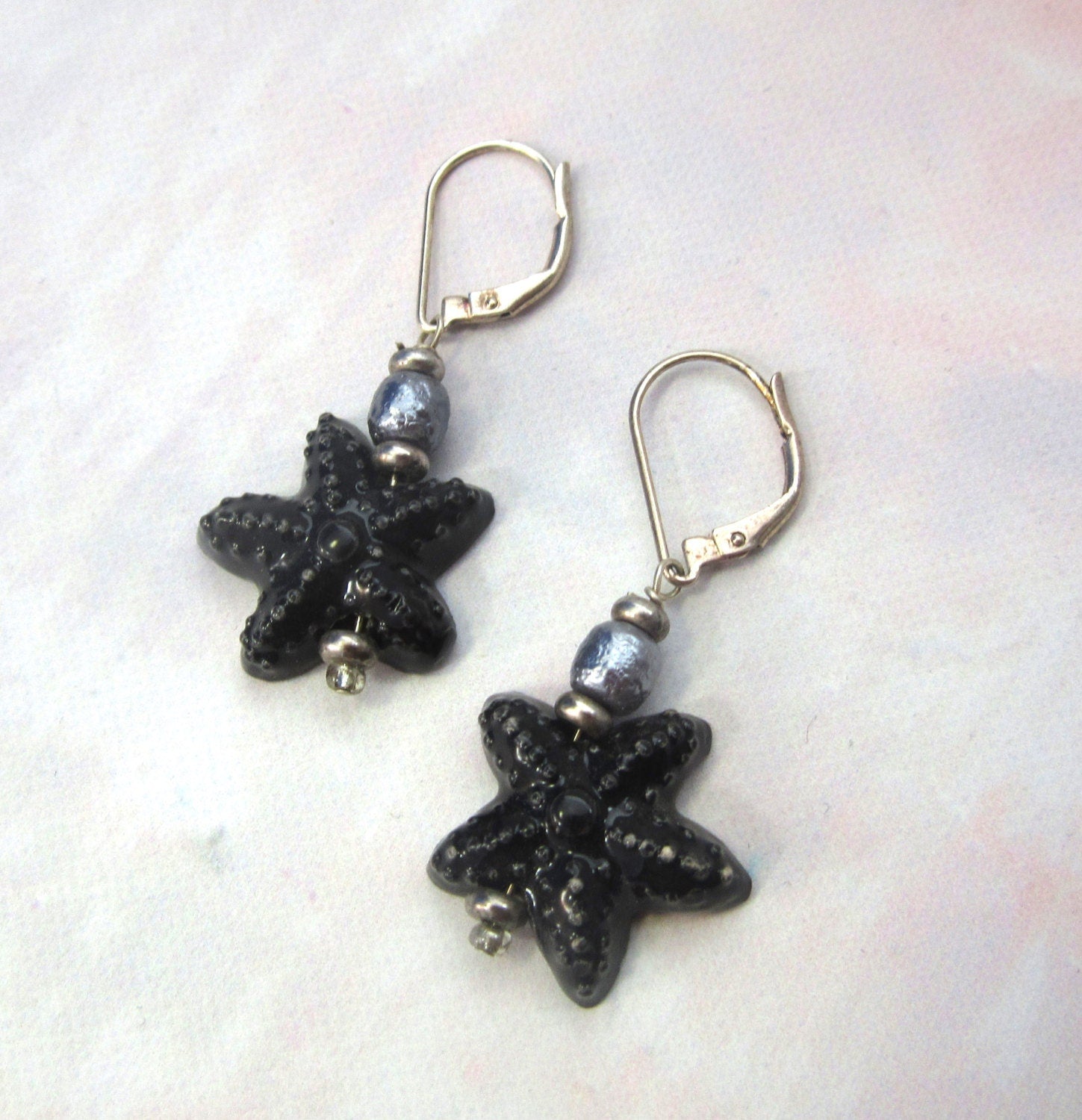 Hand-Painted Starfish Earrings feature sterling silver, freshwater pearl toppers, and intricate starfish designs. Displayed on a light background.