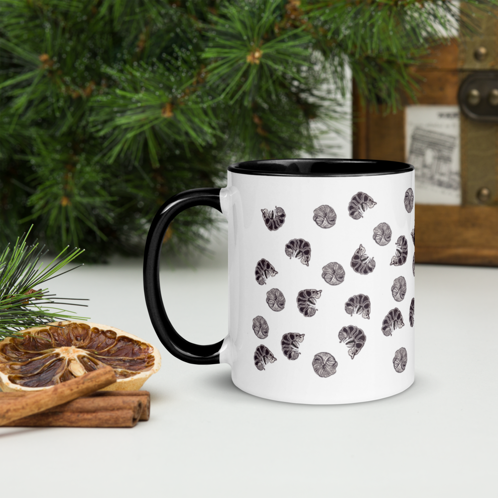 A Roly Poly Pill Bug Isopod Mug with Color Inside from Enlivenadorn by Gwynstone, featuring a white exterior, black handle, and black pill bug design, sits on a white surface near pine branches, dried citrus slices, cinnamon sticks, and a vintage wooden box in the background.