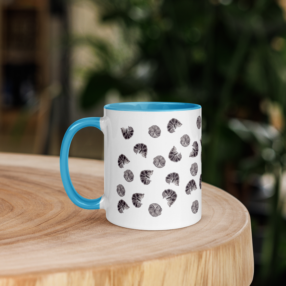 A Roly Poly Pill Bug Isopod Mug with Color Inside by Enlivenadorn by Gwynstone––white with a blue handle and rim, and adorned with a playful pattern of small black pill bug illustrations––rests on a light wooden table against a softly blurred green backdrop, making it the perfect mug for any arthropod enthusiast.